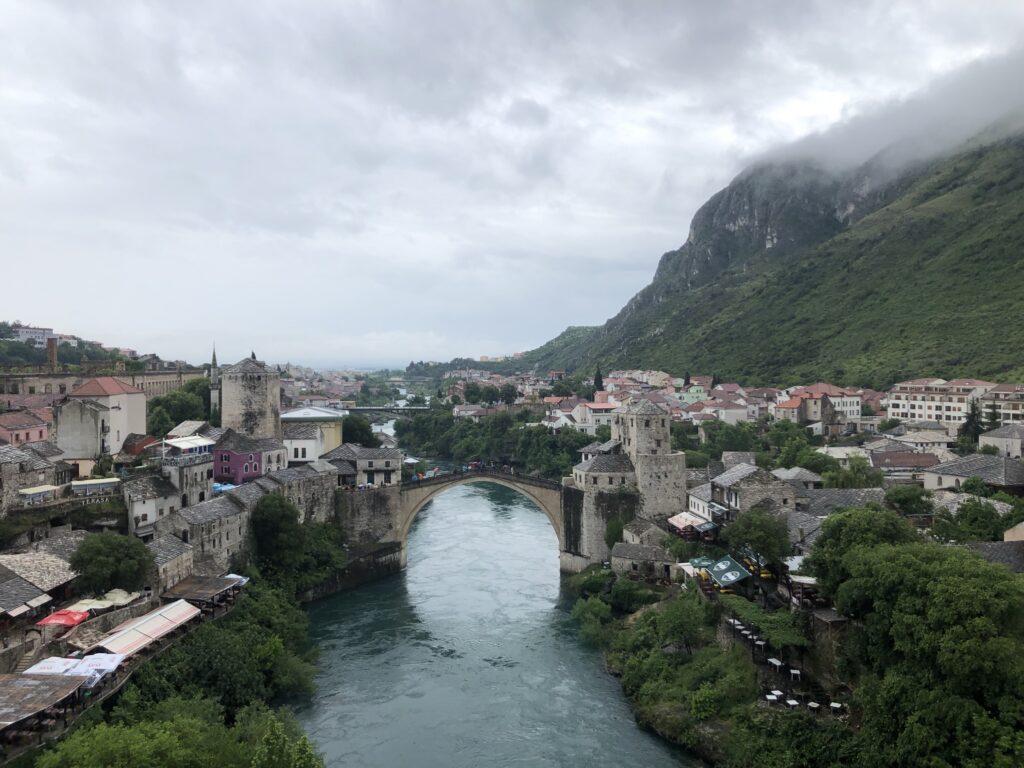 A scenic view of the Old Bridge in Mostar, Bosnia, spanning over the Neretva River, surrounded by historical buildings and lush green hills under a cloudy sky.