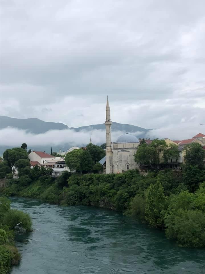 A view of a river flowing beside a mosque with a tall minaret, surrounded by greenery and cloudy mountains in the background.