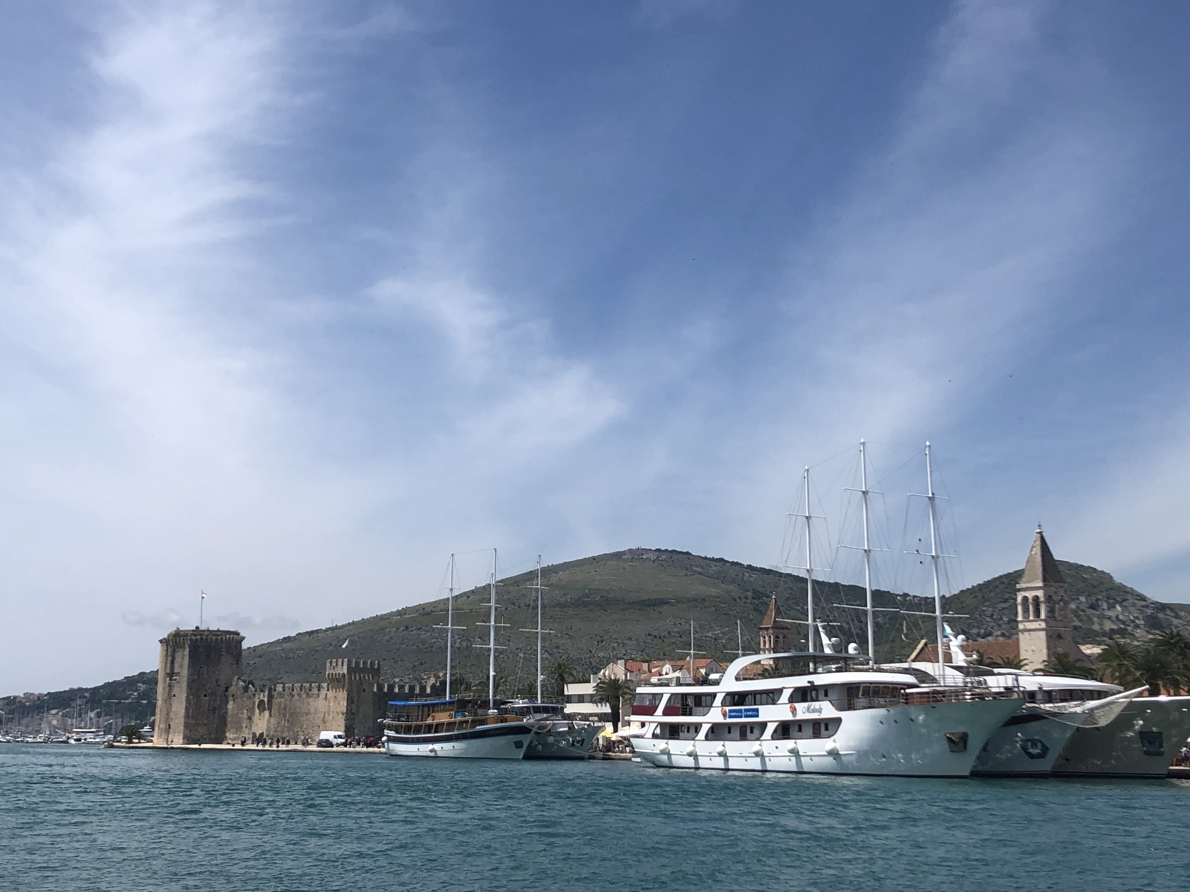 A scenic view of a marina featuring several boats anchored near a rocky fortress and a mountain, under a partly cloudy sky.