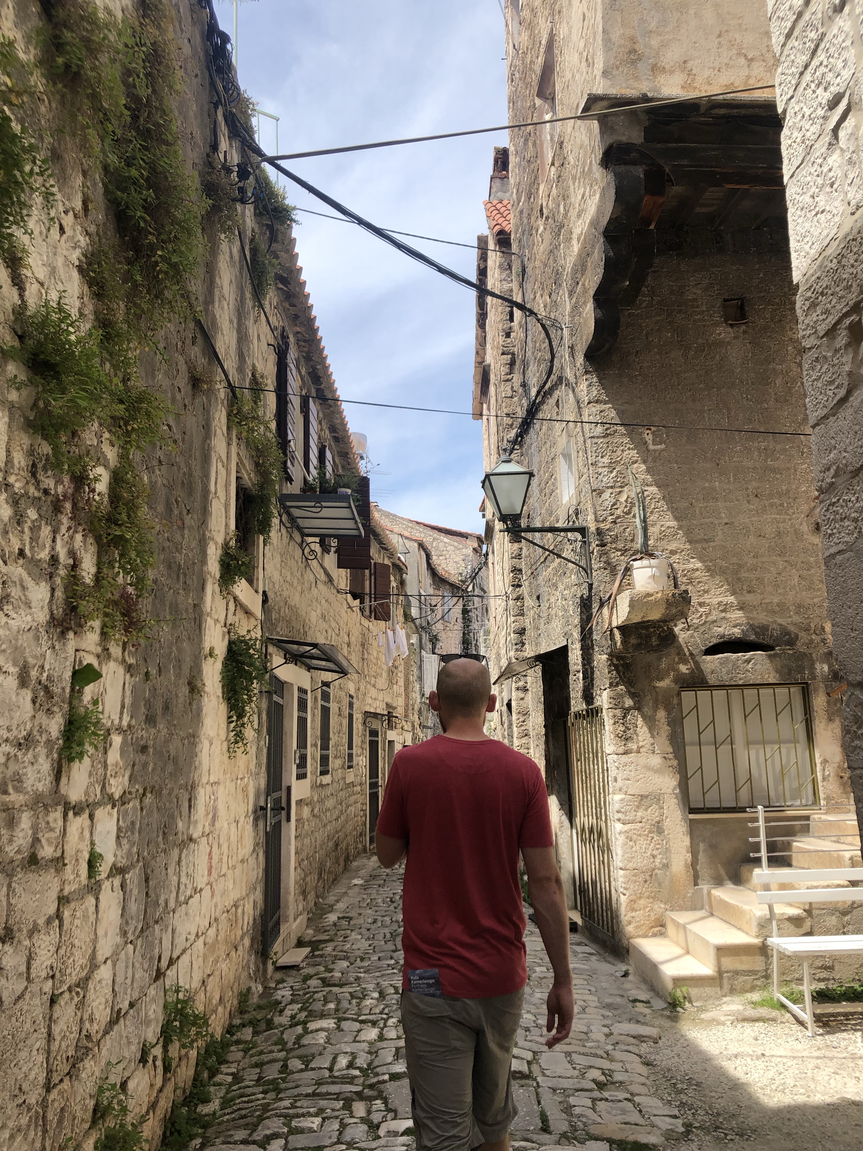 A person walking through a narrow, cobblestone alleyway lined with historic stone buildings, some featuring greenery and hanging laundry.