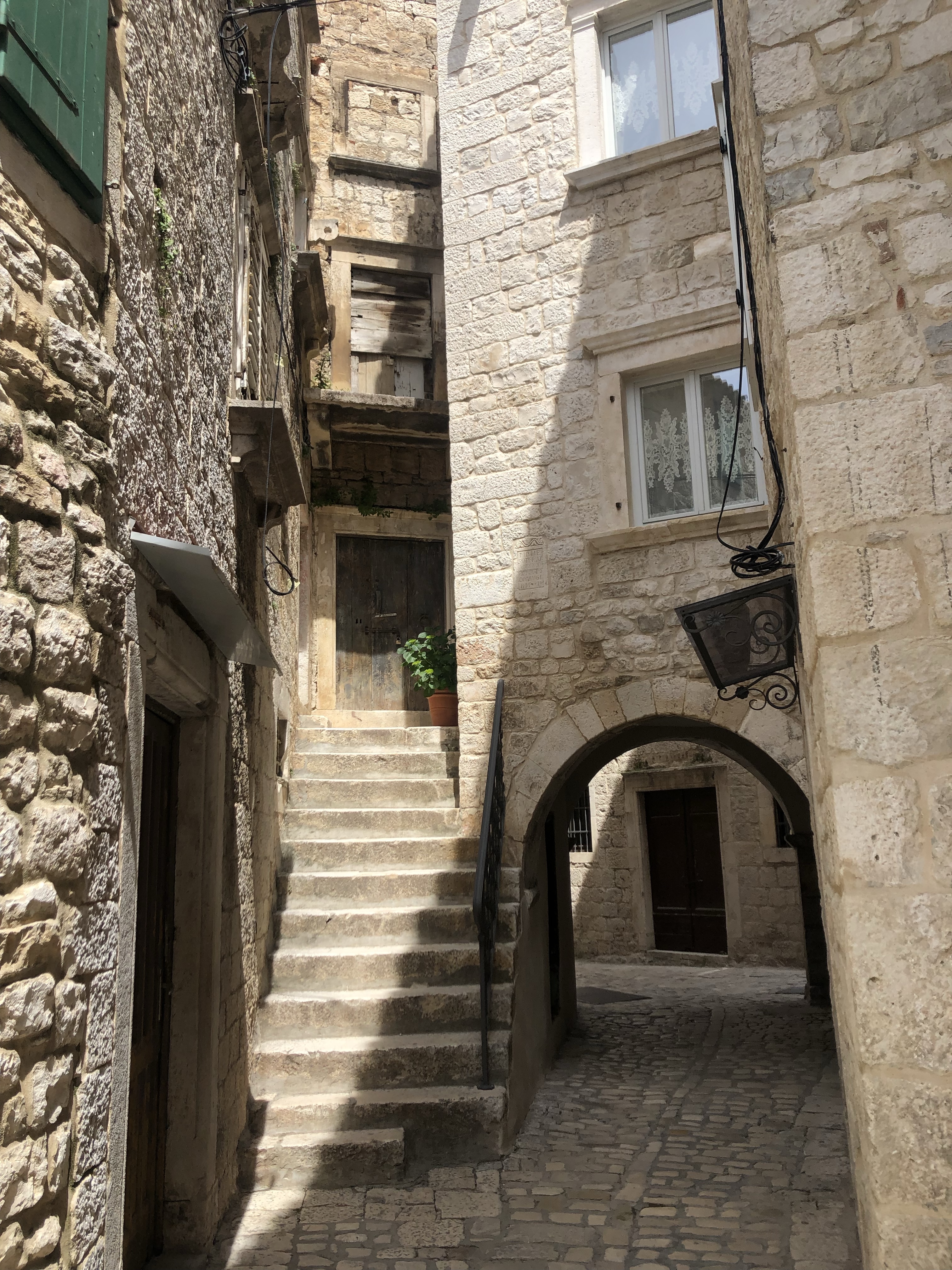 Narrow alleyway with cobblestone pavement, featuring stone buildings and a set of stairs leading to an entrance, illuminated by natural light.