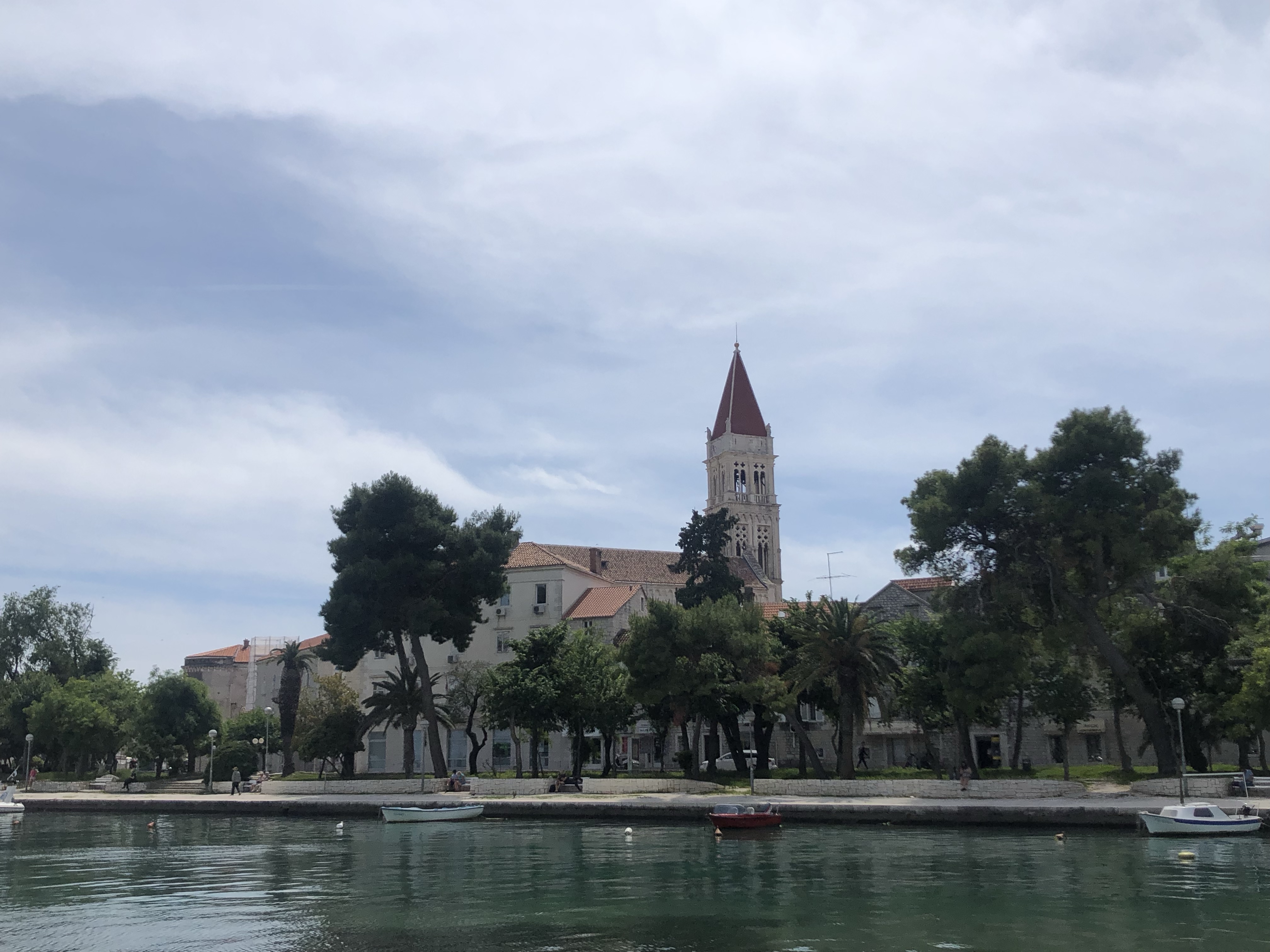 View of a coastal town with a church tower featuring a red spire, surrounded by palm and pine trees, and small boats docked along the waterfront.