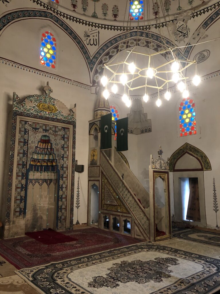 Interior of a mosque showcasing colorful stained glass windows, an ornate prayer niche, and a staircase leading to a platform. The walls are decorated with intricate patterns and calligraphy, while a modern chandelier hangs from the ceiling.