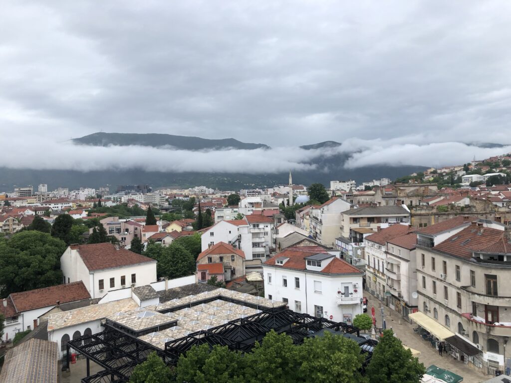 A view of a cityscape with red-tiled roofs and a backdrop of mountains partially covered by clouds, under a cloudy sky.