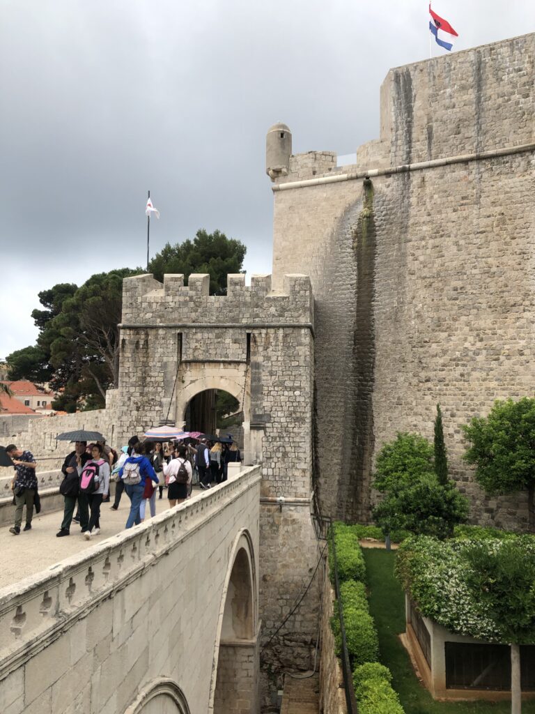 A historic stone fortification with an archway, featuring tourists walking across a bridge. The scene includes green vegetation and a cloudy sky.