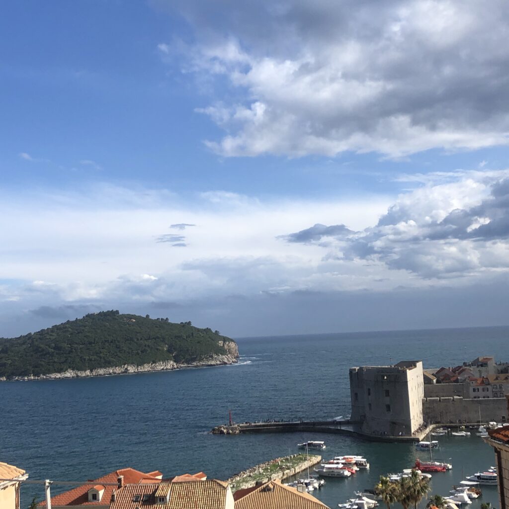 Scenic view of a coastal landscape featuring an island, calm sea, and cloudy sky, with boats docked at a harbor and historical buildings along the shore.