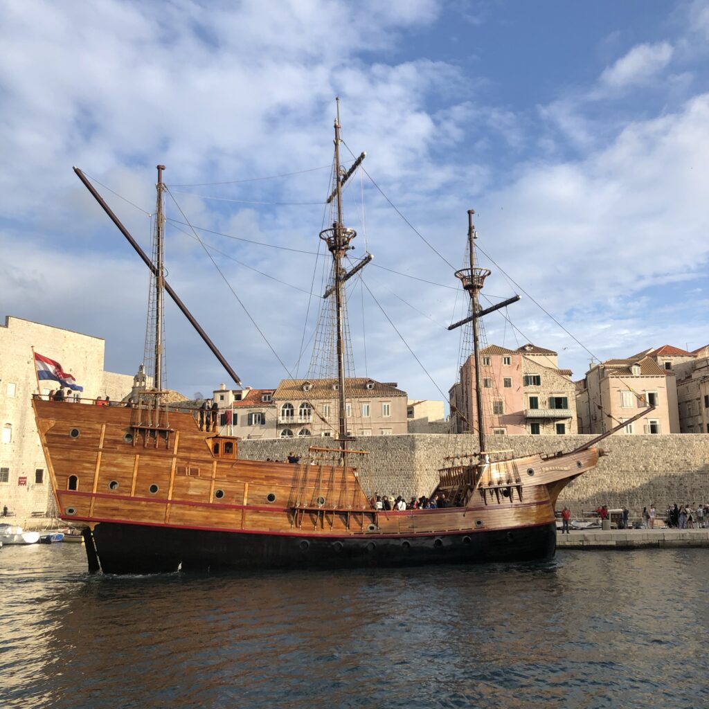 A large wooden sailing ship docked at a harbor with historic buildings in the background and blue skies above.