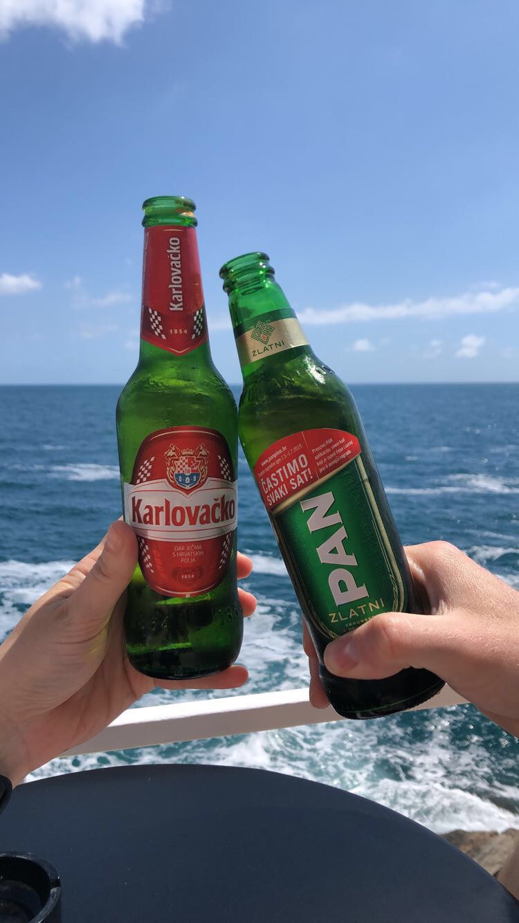 Two hands clinking bottles of Karlovačko and Pan beer against a backdrop of the ocean under a clear blue sky.