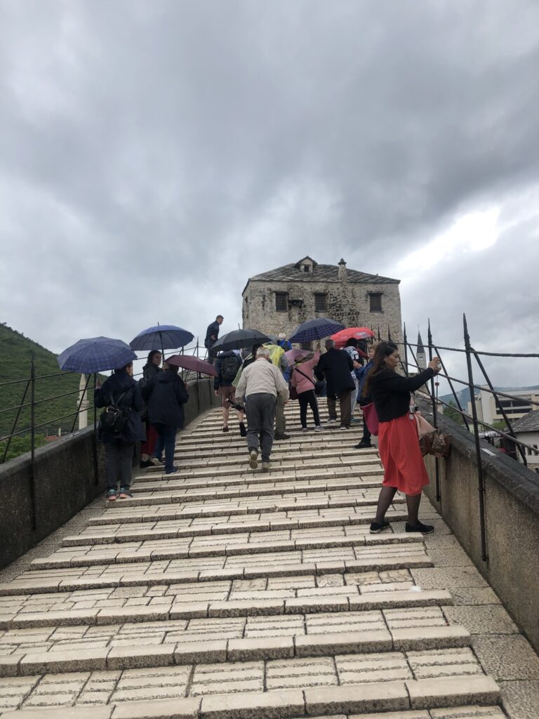 Walking across the steep Stari Most Bridge with many other people holding umbrellas.