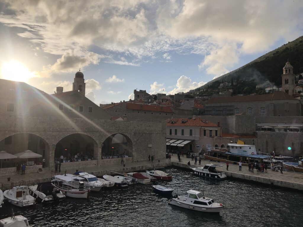 A scenic view of a harbor with several boats docked, surrounded by historic stone buildings and red-tiled roofs, under a partially cloudy sky with the sun shining through.