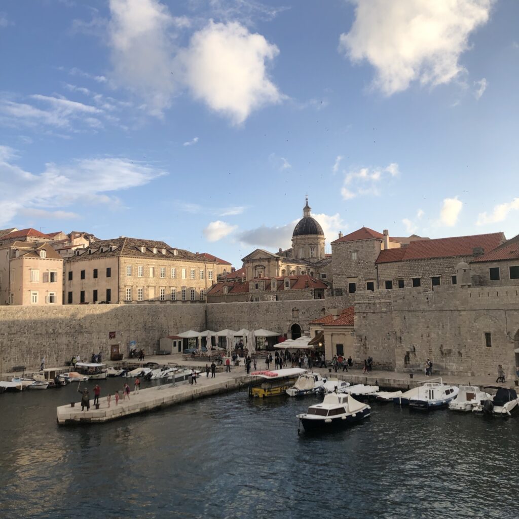 A scenic view of a harbor surrounded by historic buildings with terracotta roofs and a prominent dome, under a partly cloudy sky.