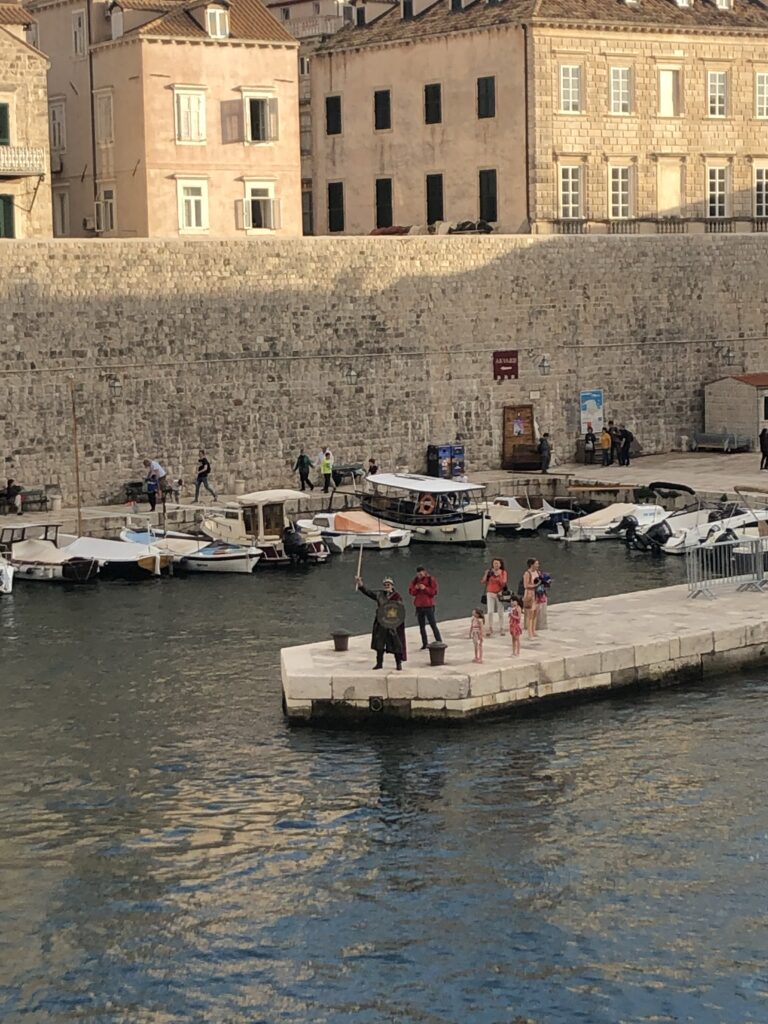 A group of people standing on a dock by a harbor, with boats in the water and a stone wall in the background. One person is holding a faux sword and wearing a costume.