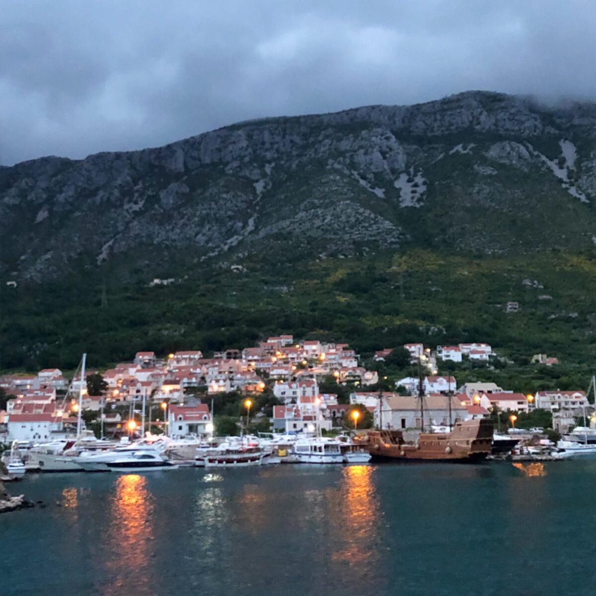 A scenic view of a coastal town at dusk, featuring a harbor filled with boats and a large wooden ship. The background includes green hills and a cloudy sky, with lights from the town reflecting on the water.
