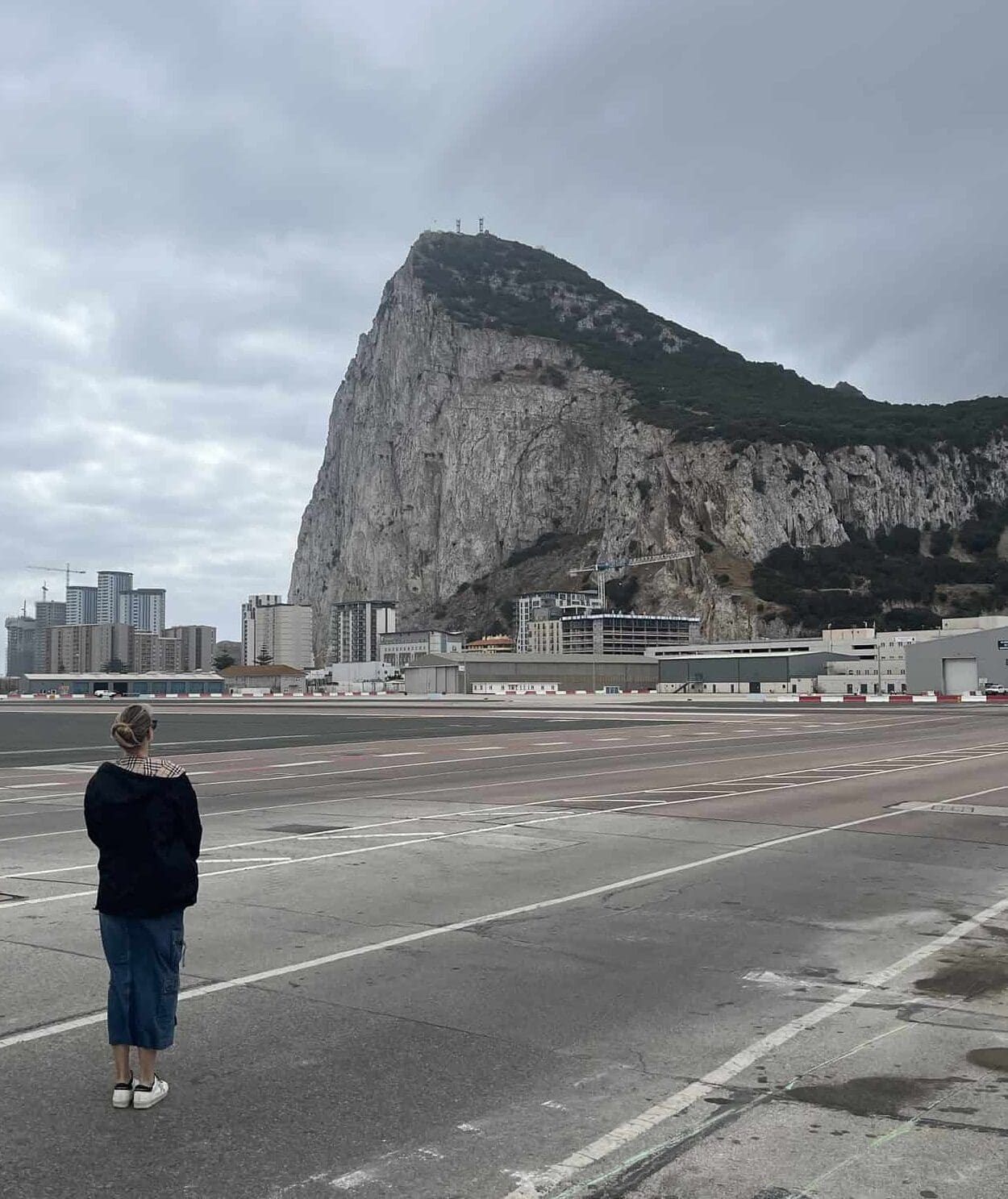 Woman standing on Gibraltar's famous airport runway, looking up at the Rock of Gibraltar under a clear sky.
